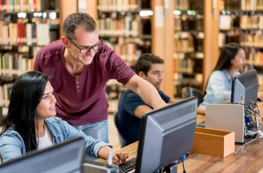A man and woman sit at computers in a library, focused on their work amidst bookshelves and study materials.